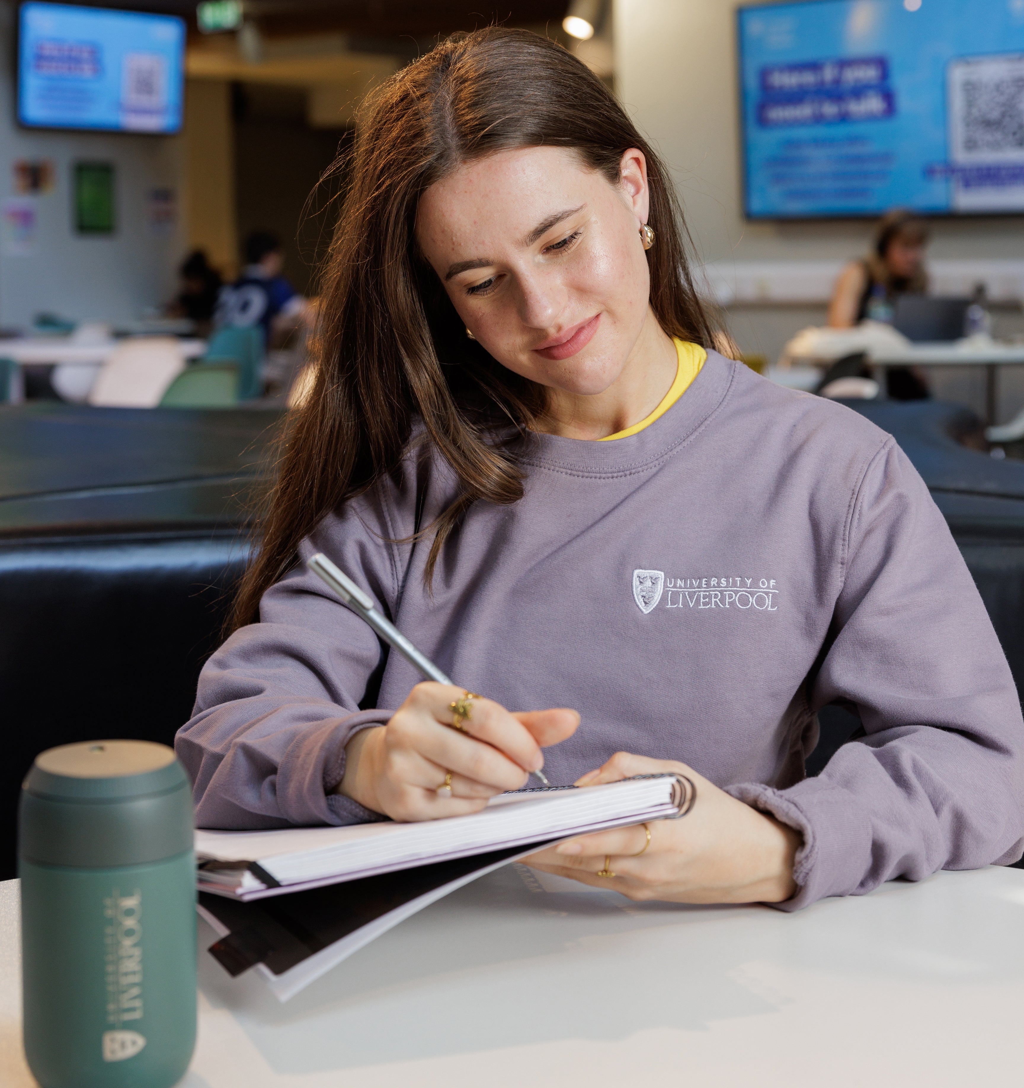 student in a University of Liverpool  purple sweatshirt with a logo, writing in a notebook at a table.